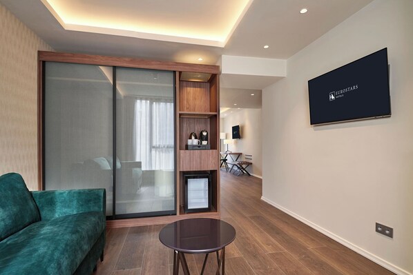 A modern hotel room with a sliding glass door, a green sofa, a small table, and a mounted screen displaying hotel branding. - Eurostars Avenue Louise (Brussels)