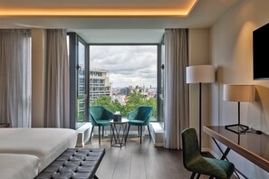 Hotelkamer met een bed, twee stoelen, een kleine tafel, een bureau met lamp en uitzicht op de stadszicht door grote ramen.
