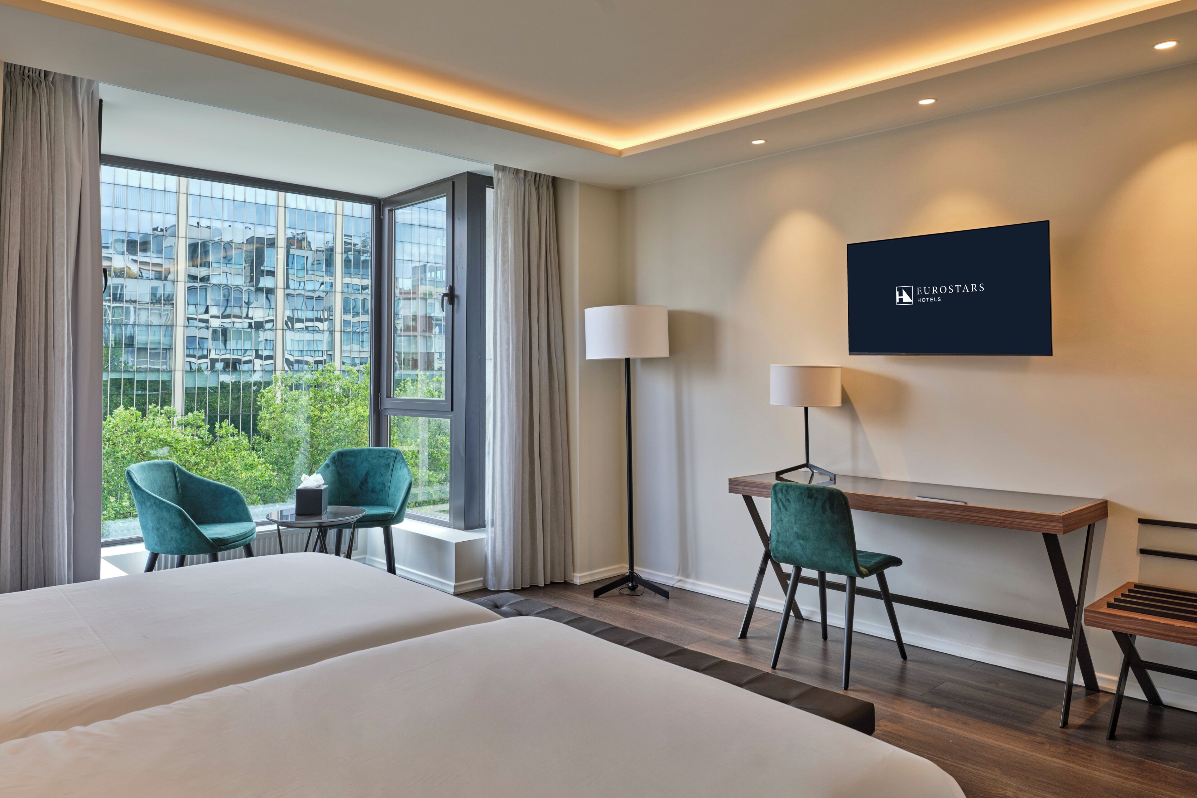 Ein Hotelzimmer mit einem Bett, zwei Stühlen, einem Schreibtisch, einem Fernseher und Blick auf eine Stadtlandschaft durch große Fenster.