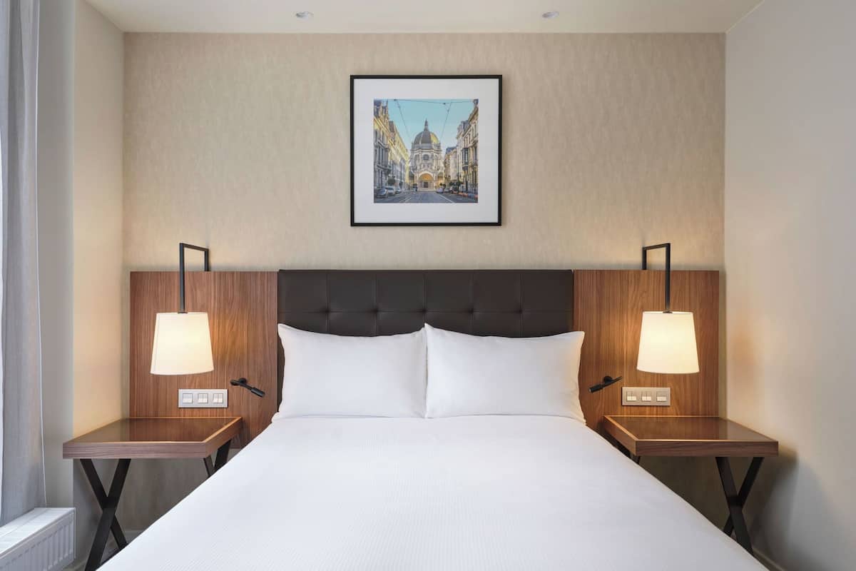 A hotel room with a large bed, two bedside tables with lamps, a framed picture on the wall, and a radiator.