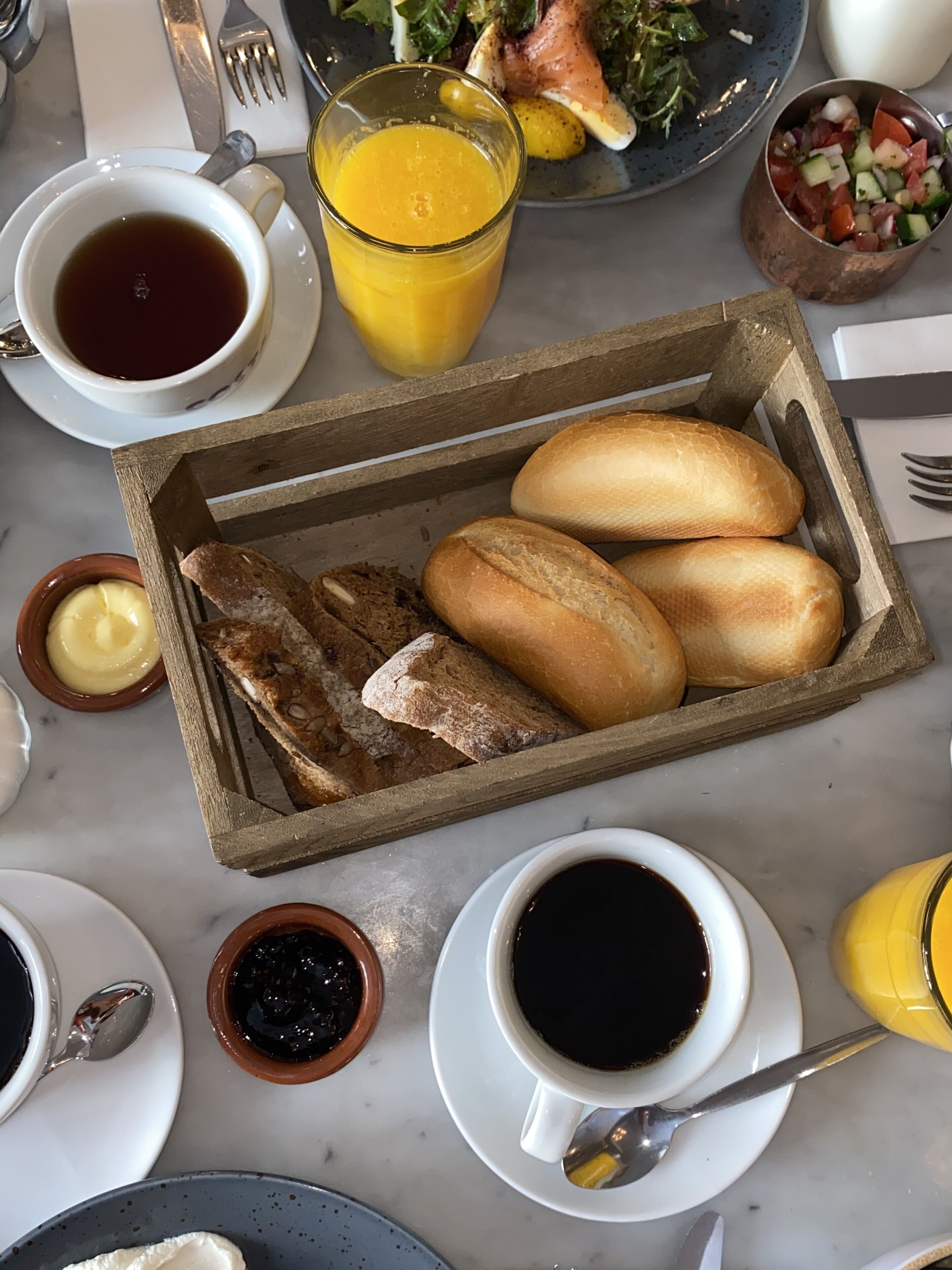 Desayuno con pan, mantequilla, mermelada, café y un jugo de fruta sobre una mesa de mármol.