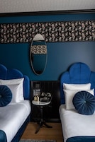 A room with two beds, each with a blue headboard and white bedding. There is a small table with a vintage radio and a decorative item. The room has a dark blue wall with a floral pattern and a mirror above the bed.