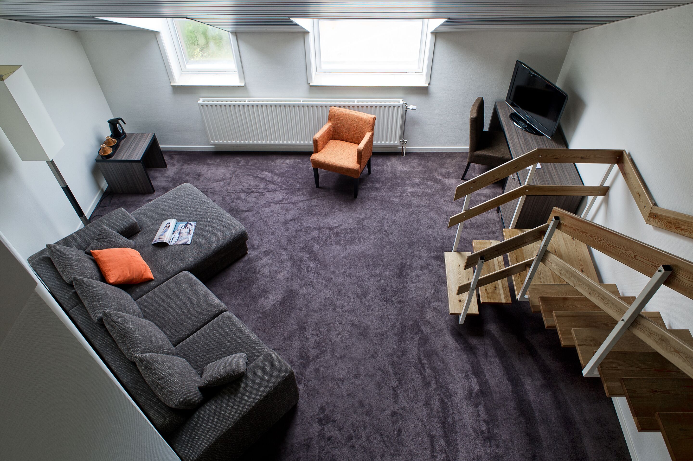 Un apartamento moderno con un sofá gris, un sillón naranja, una escalera de madera y un televisor sobre una mesa de madera.