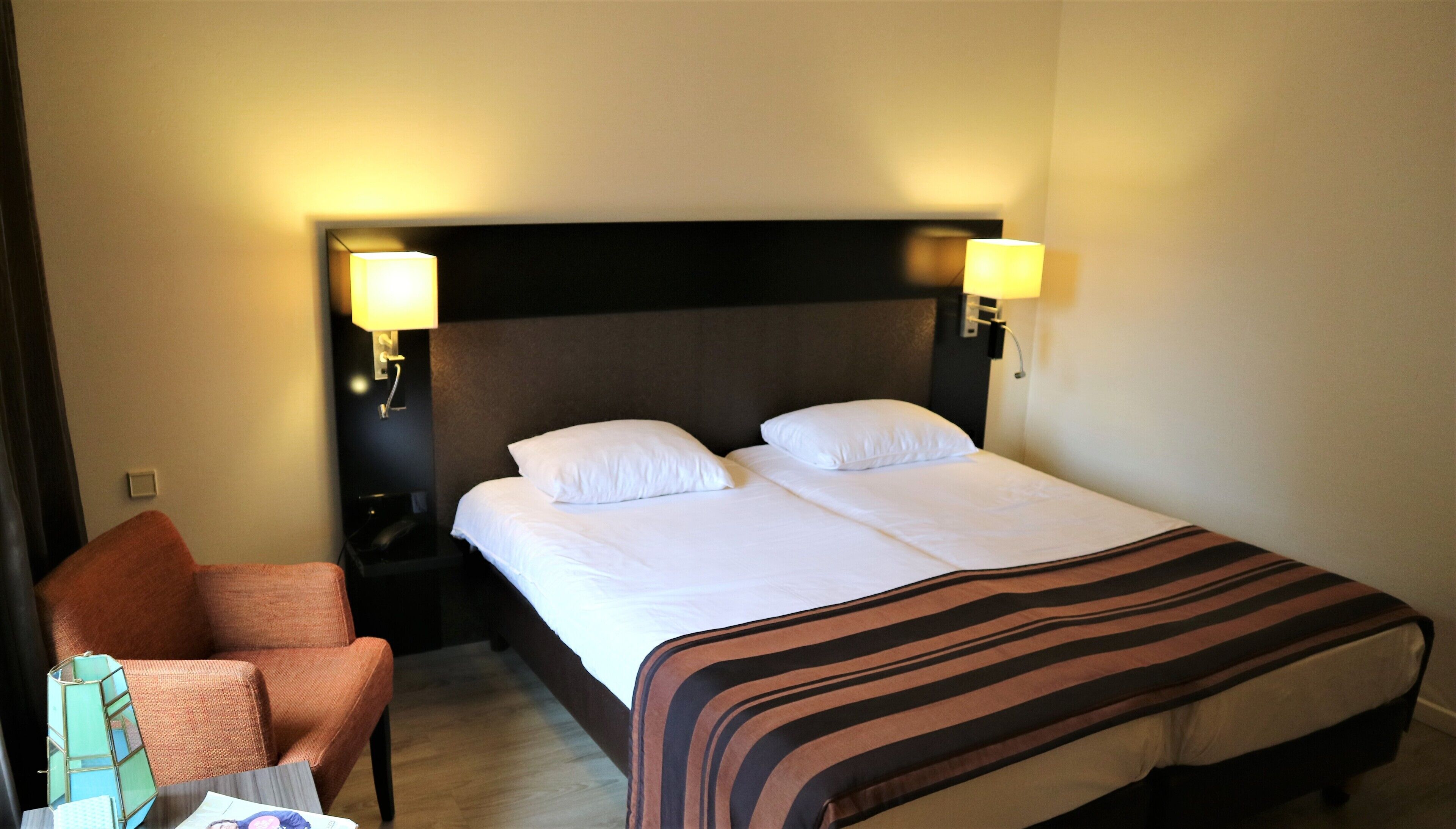 A hotel room with a double bed, two bedside lamps, a nightstand, a chair, and a striped blanket.