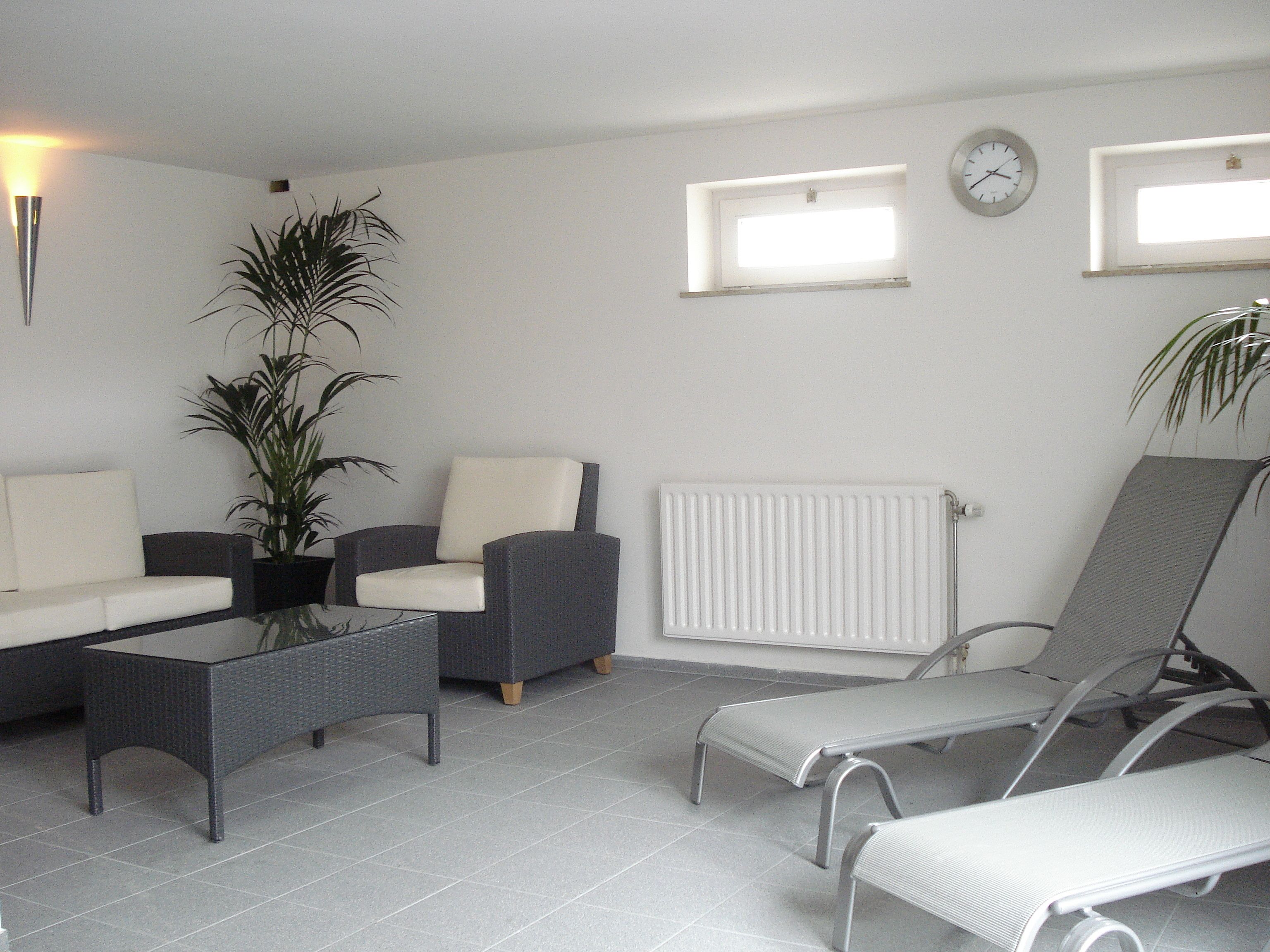 A modern living room with a sofa, armchairs, a coffee table, and a lounge chair. There are two windows with blinds, a radiator, and a clock on the wall.