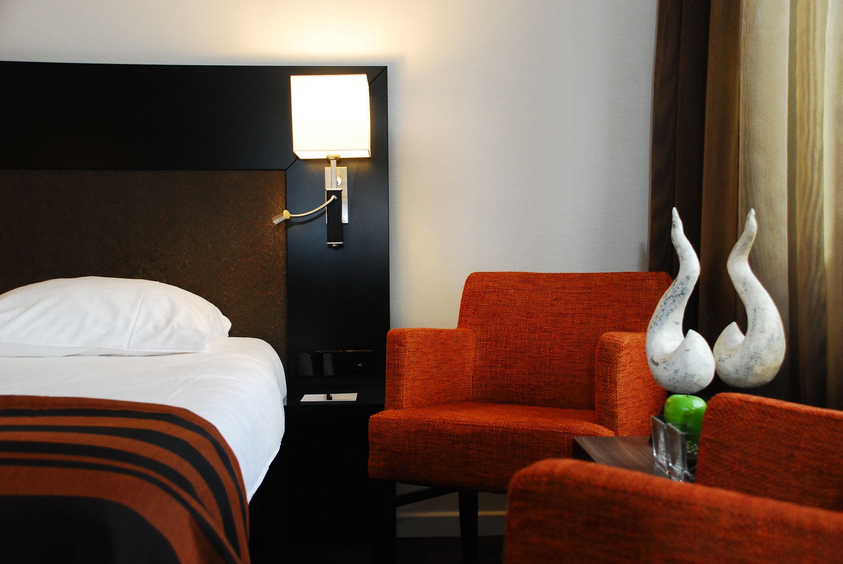 Een hotelkamer met een bed, nachtkastje, lamp, oranje stoel en een decoratief beeldhouwwerk.