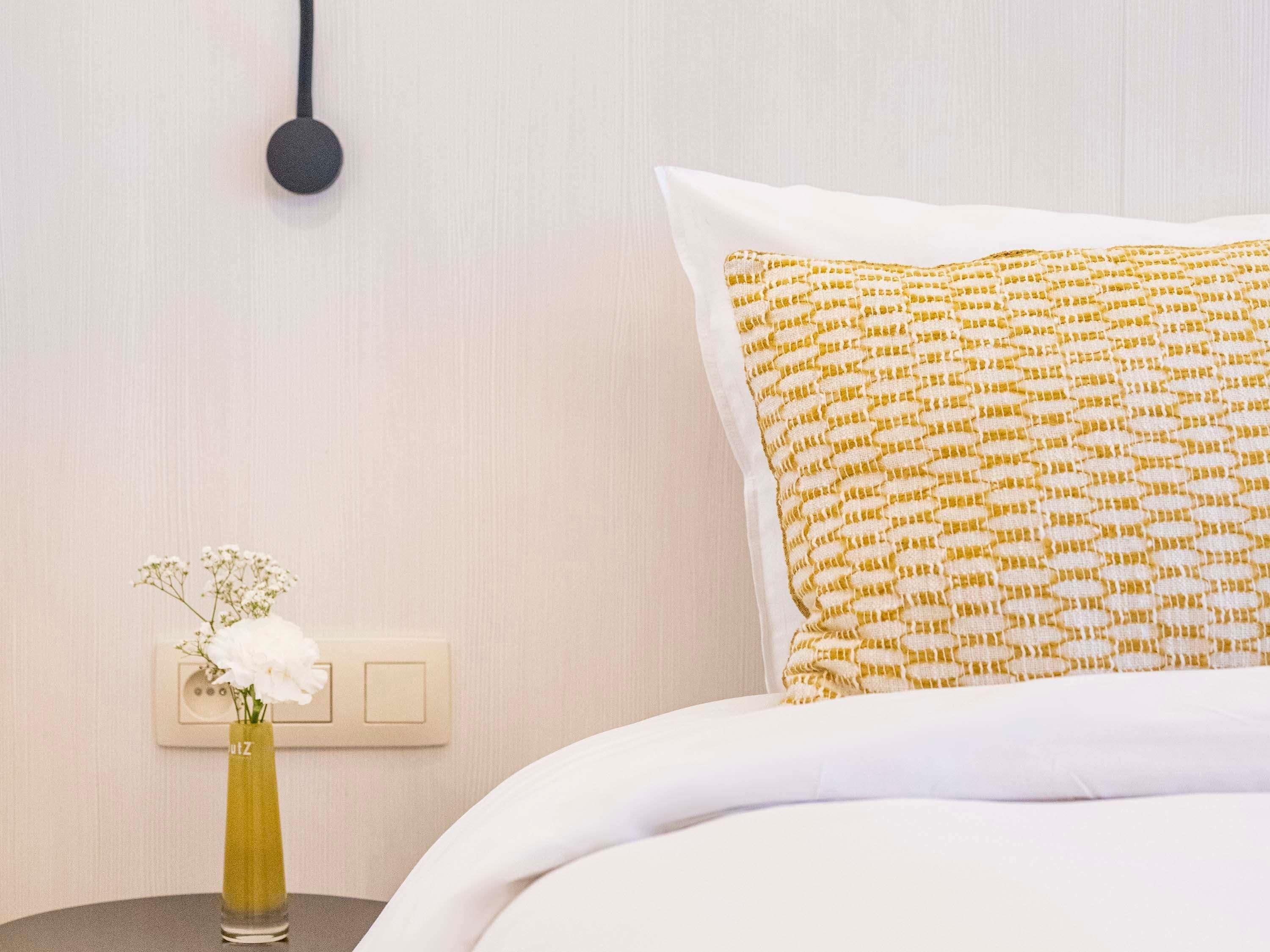 Un lit bien fait, agrémenté d’un coussin à motifs, une table de chevet avec un vase de fleurs et un luminaire fixé au mur.