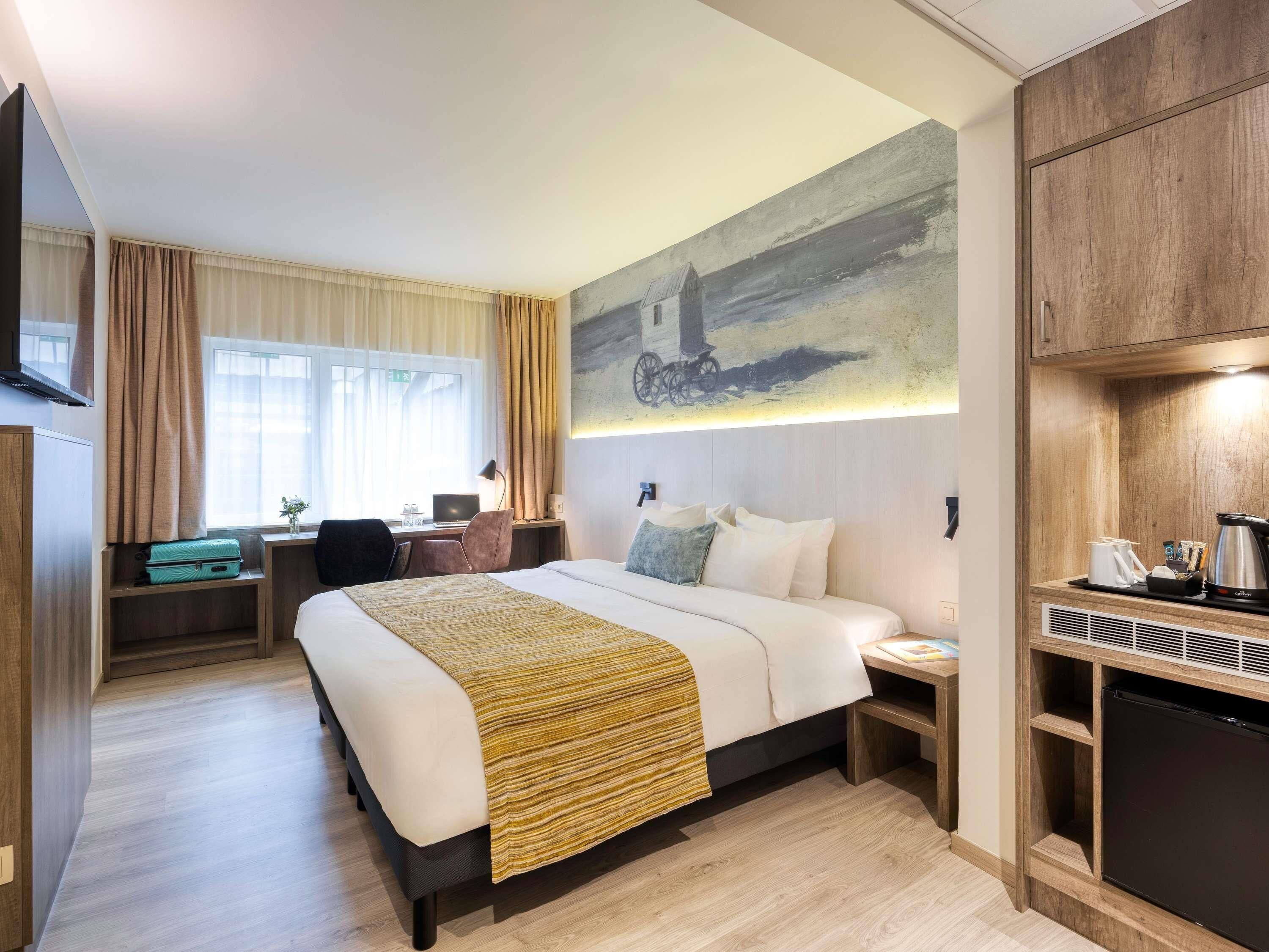 A hotel room with a bed, desk, chair, and a painting on the wall.