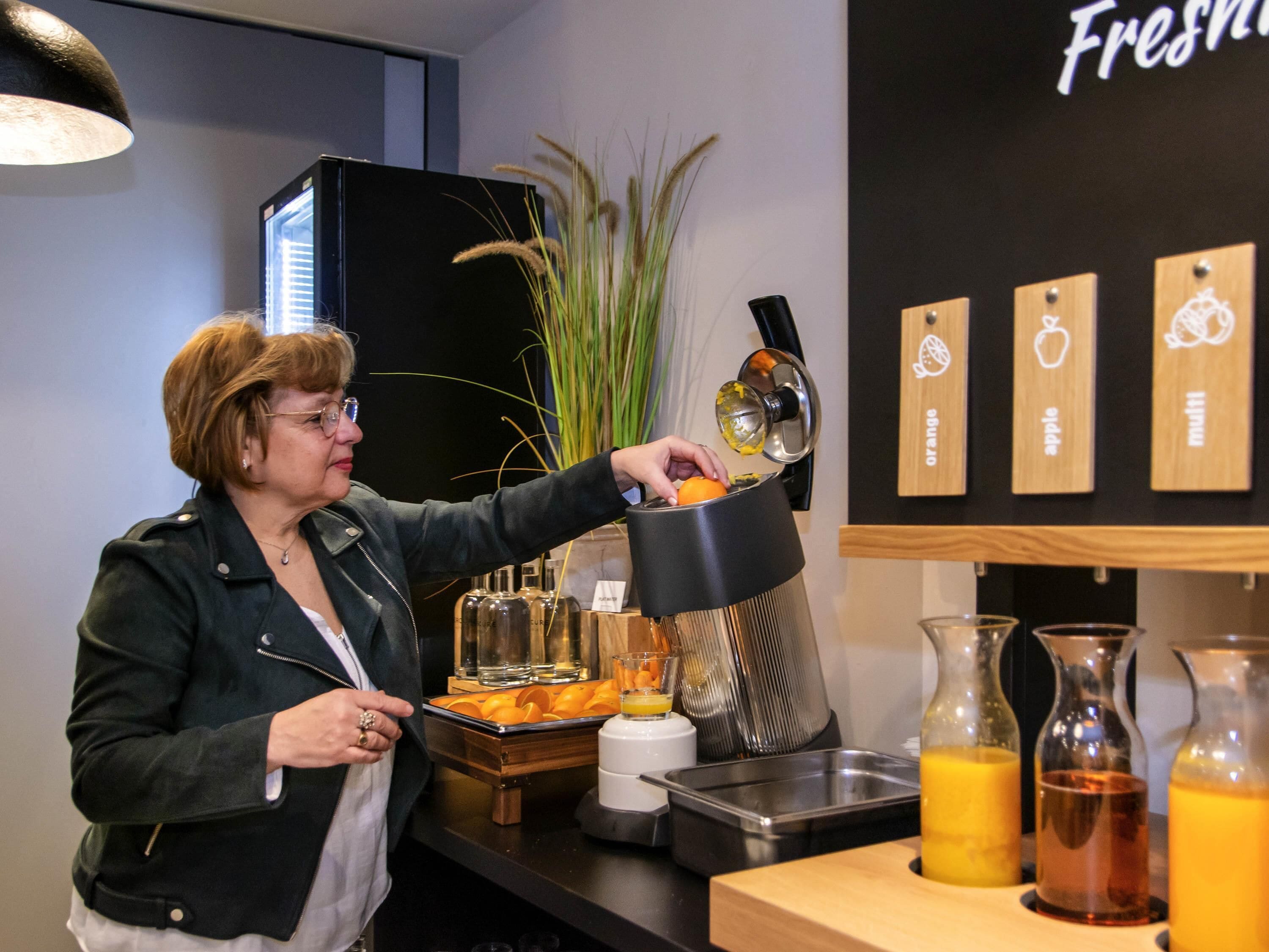 A person juicing oranges at a kitchen counter with a juicer, a sink, and a shelf with labeled containers.