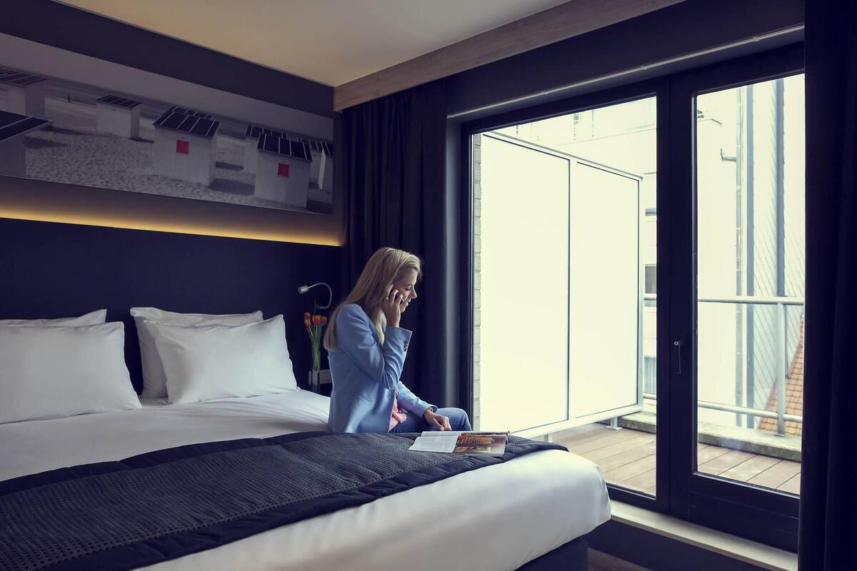 A modern hotel room with a large bed, a desk, and a view of the city through a large window.