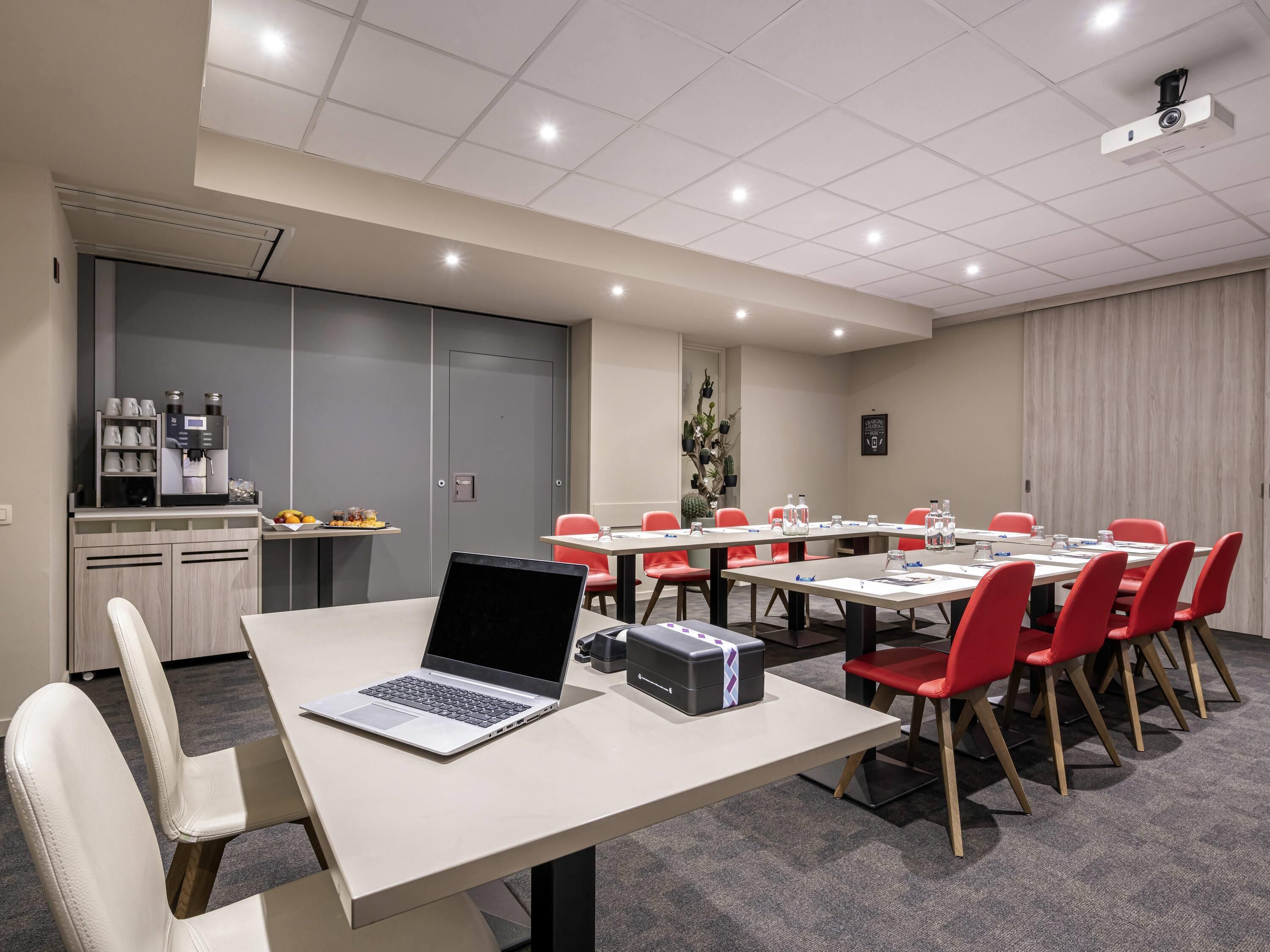 Un salón de conferencias con una larga mesa, sillas rojas, una computadora portátil, un proyector y una máquina de café.