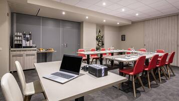 Un salón de conferencias con una larga mesa, sillas rojas, una computadora portátil, un proyector y una máquina de café.