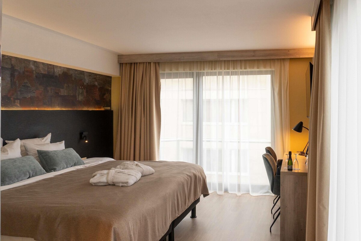 A hotel room with a bed, a desk, a chair, a painting, and a large window with curtains.