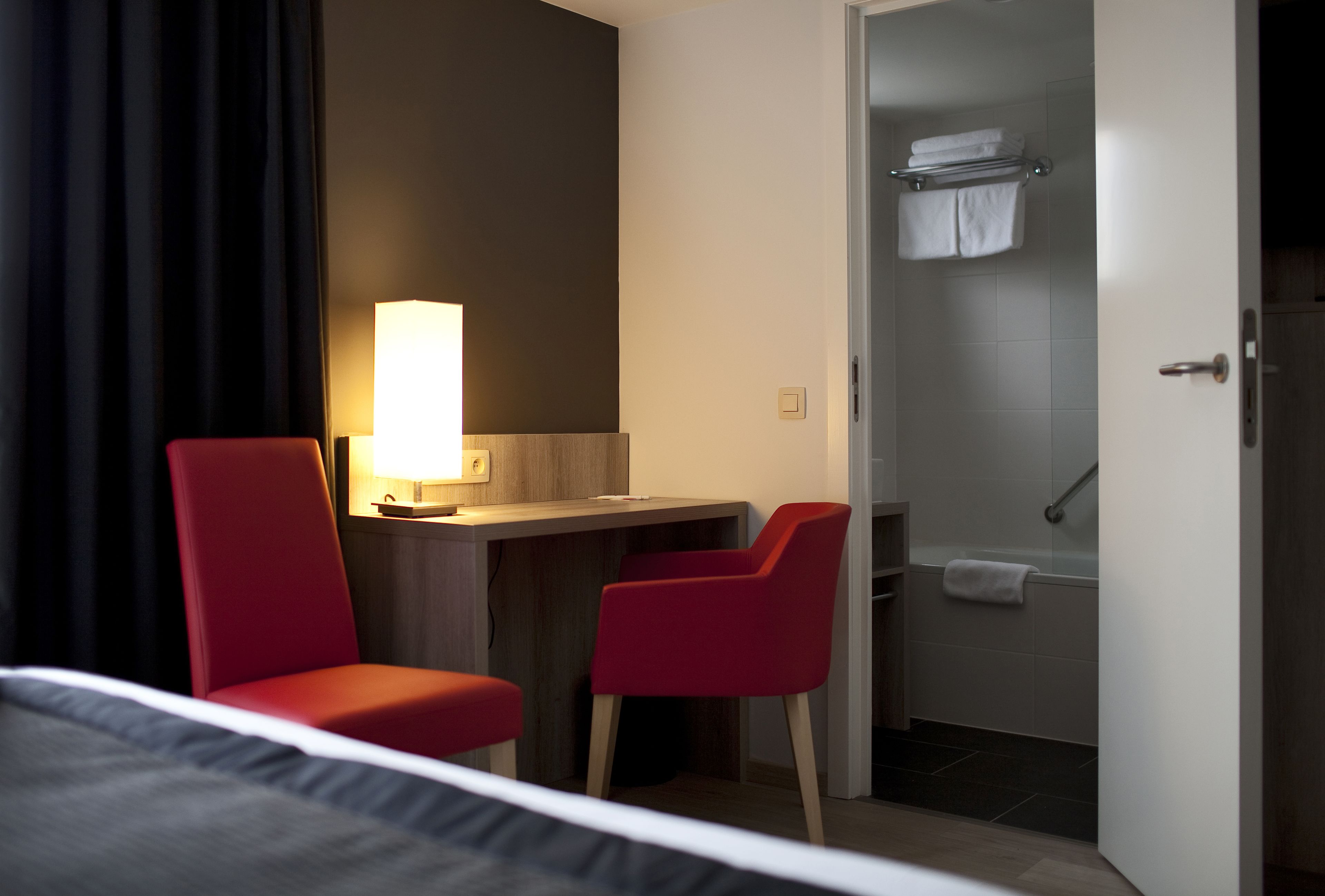 A hotel room with a bed, a desk with a lamp, two red chairs, and a shower area.