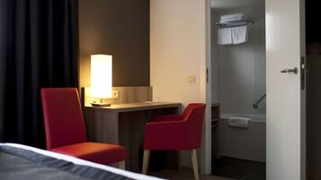 A hotel room with a bed, a desk with a lamp, two red chairs, and a shower area.