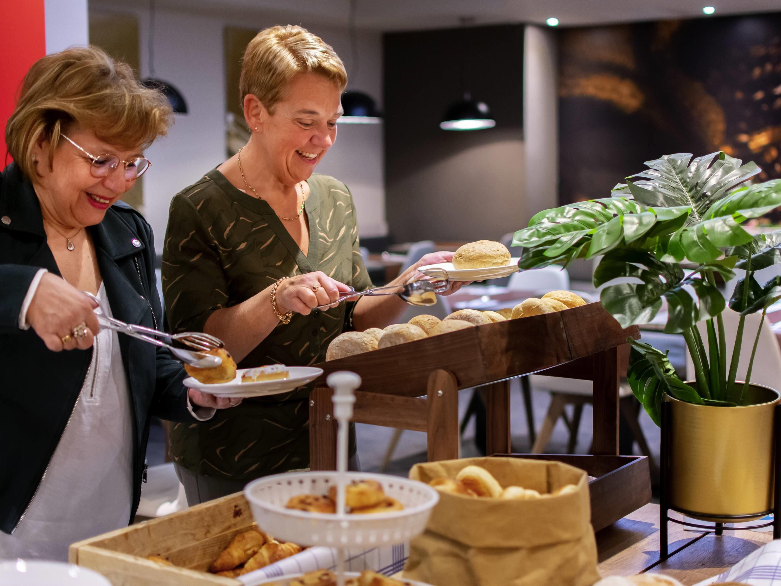 Two people serving themselves from a buffet table with various baked goods and a potted plant.