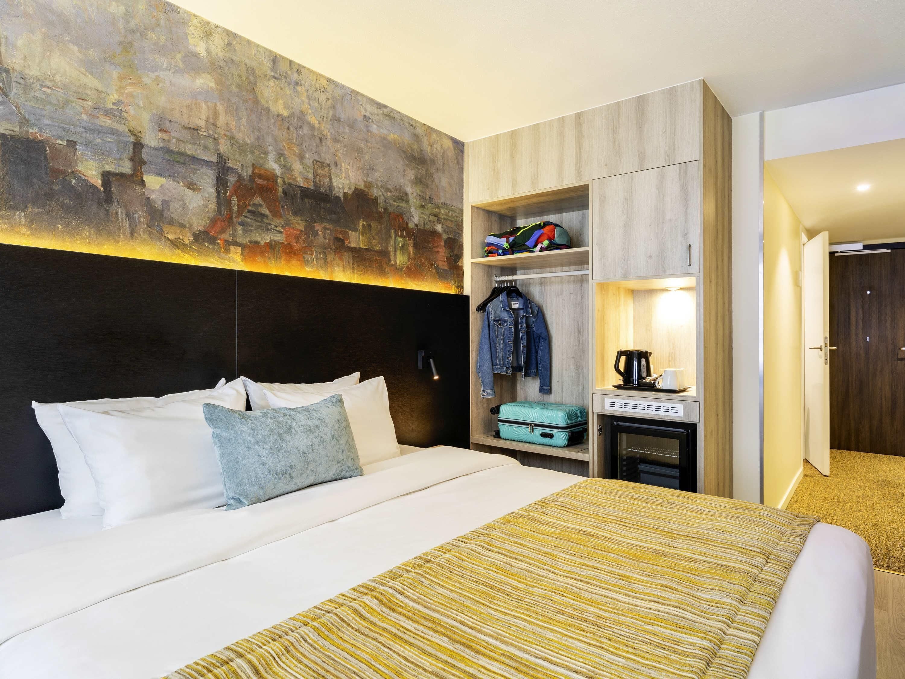 A hotel room with a large bed, a wardrobe, a small table with a kettle, and a painting on the wall.