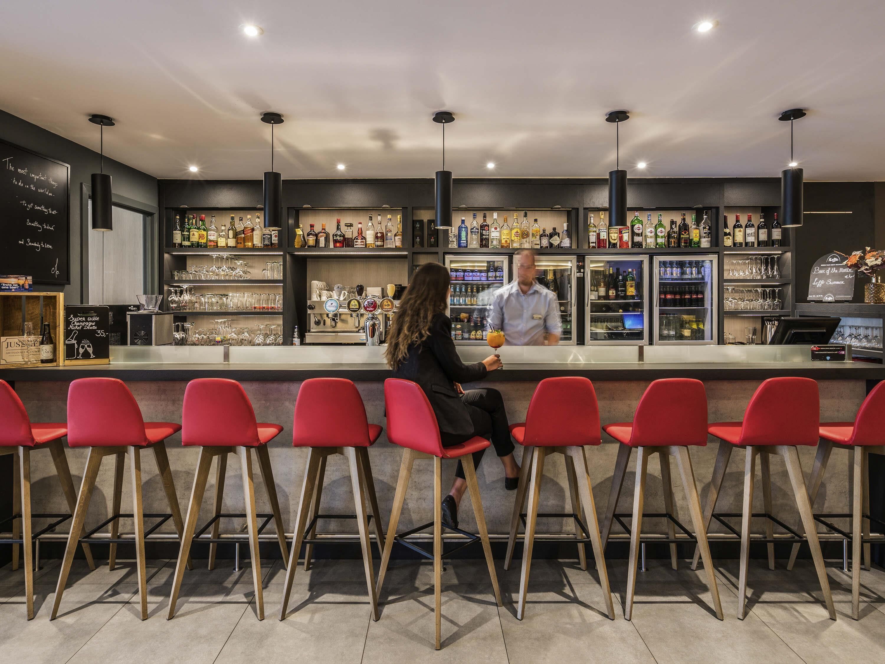 Zona de bar con sillas rojas, un barman y estantes llenos de botellas y copas.
