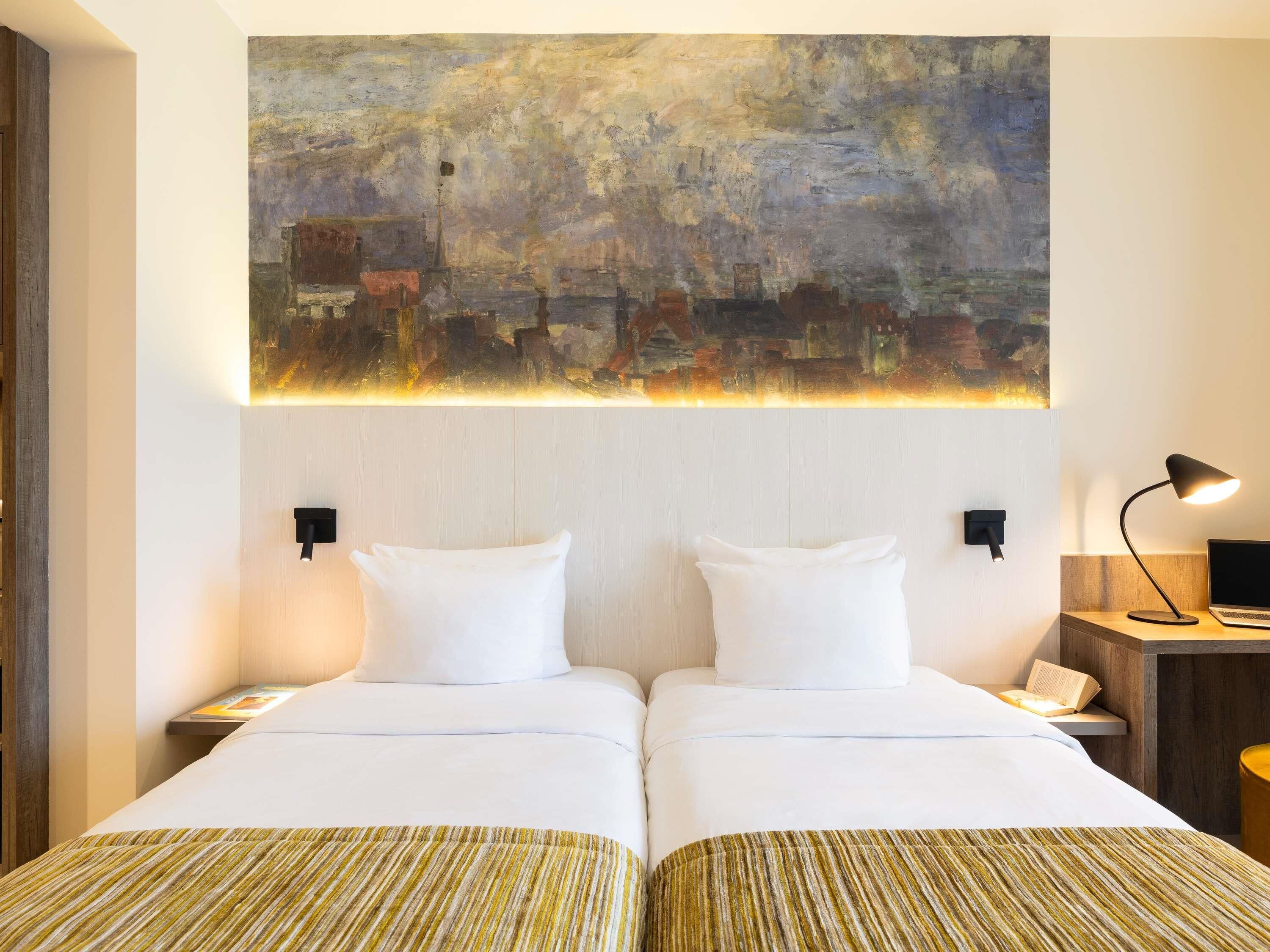 Een hotelkamer met twee bedden, een groot abstract schilderij aan de muur en een bureau met een lamp en een laptop.
