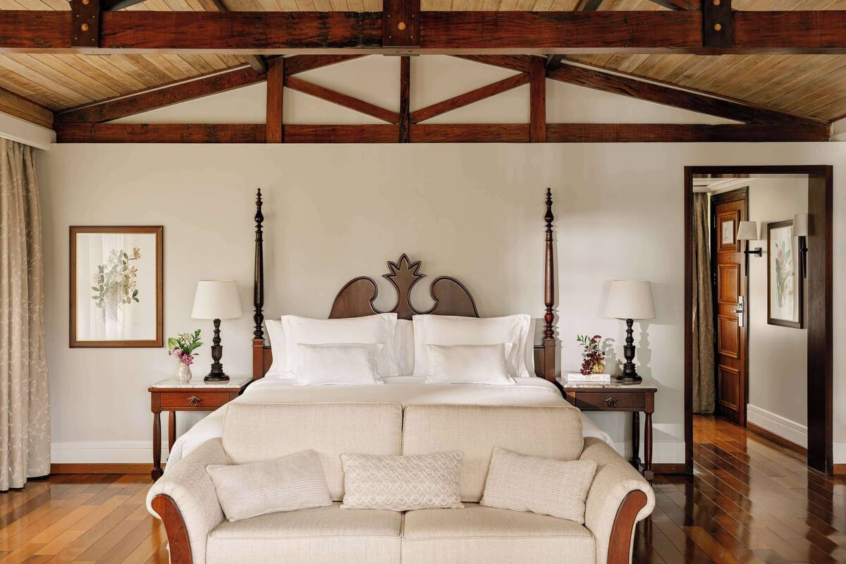 A spacious bedroom with a large bed, wooden beams on the ceiling, a sofa, and two bedside tables with lamps.