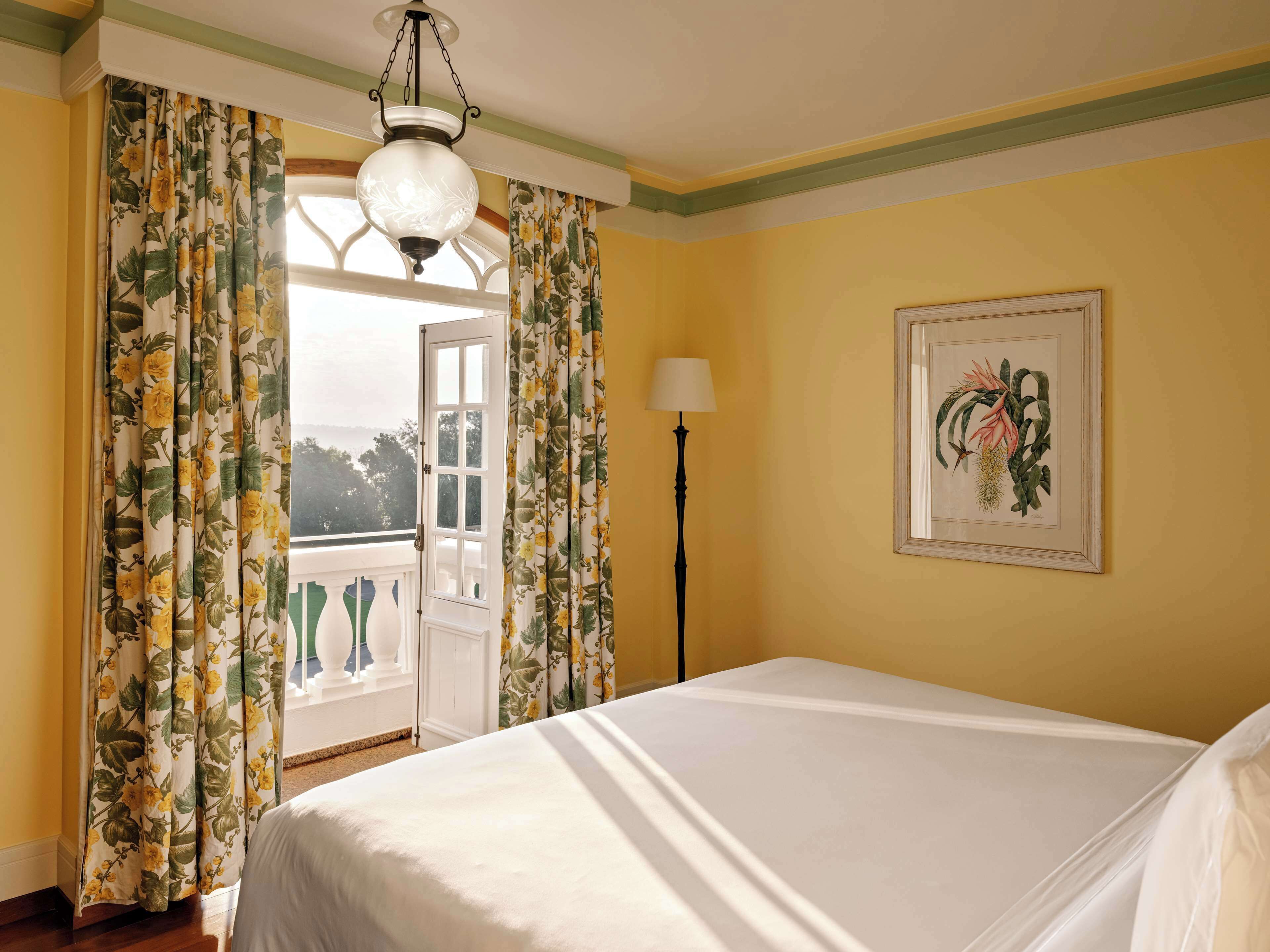 Un dormitorio con cama, cortinas, lámpara, un grabado botánico enmarco y vista a través de una puerta abierta.