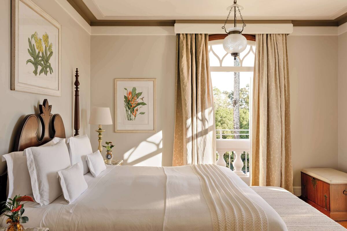 A well-lit bedroom with a large bed, a bedside table with a lamp, and framed botanical prints on the wall.