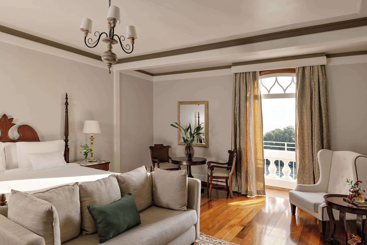 A hotel room with a bed, a sofa, a dining table, and a chair. There is a window with curtains and a view of the outside.