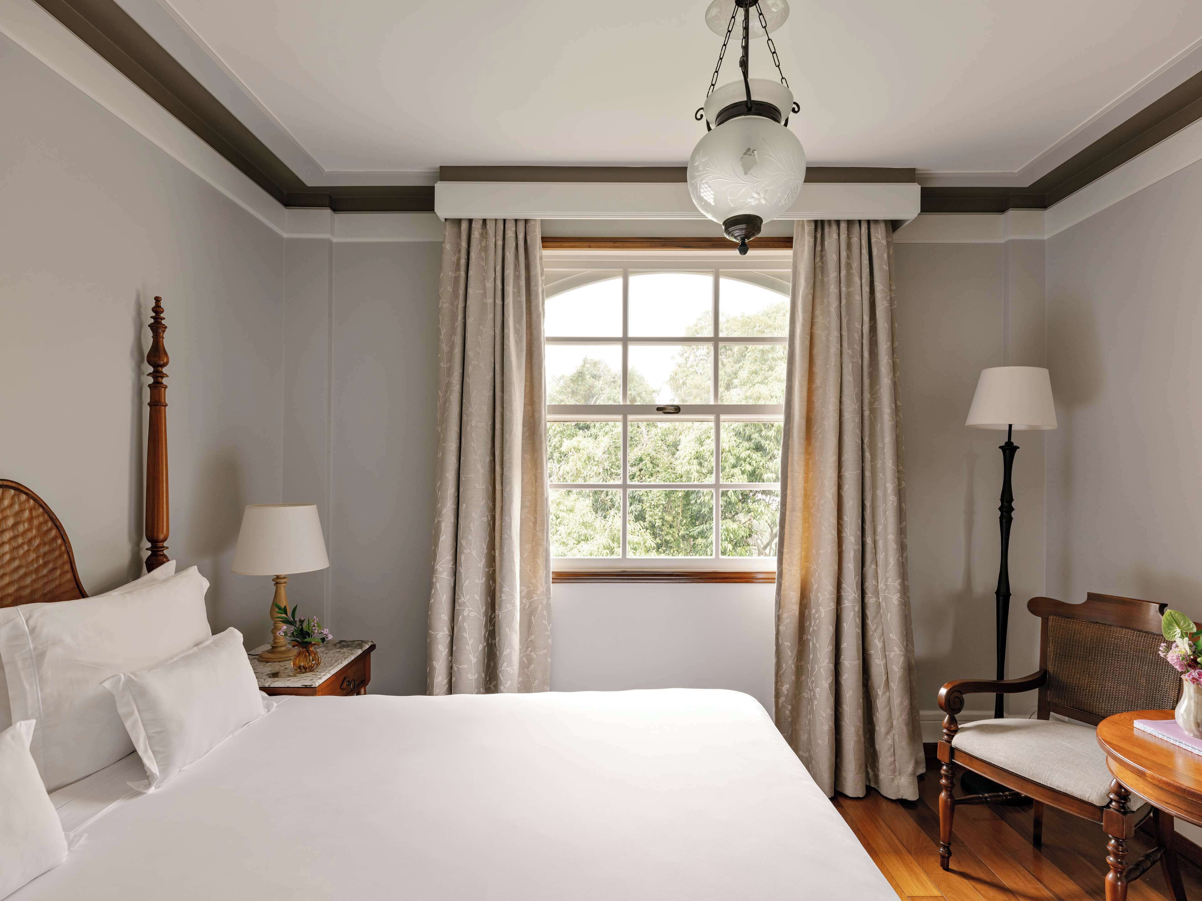 Habitación de hotel con una cama grande, una silla de madera, una mesita con lámpara y un ventanal con cortinas.
