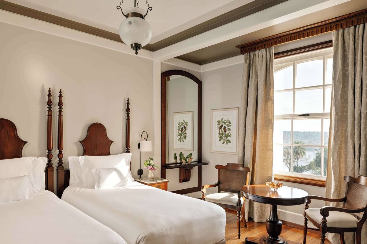 A hotel room with two beds, a wooden floor, a round table with chairs, a large window with curtains, and framed pictures on the wall.