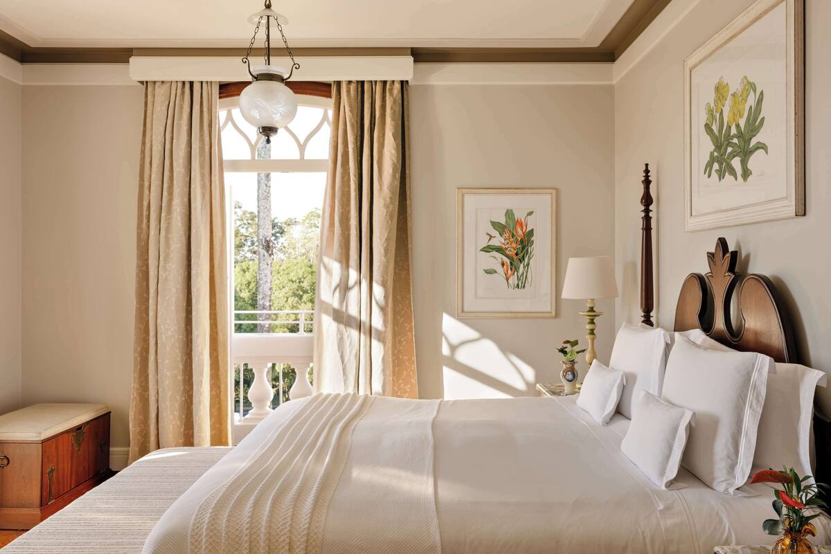 A well-lit bedroom with a large bed, a nightstand, a lamp, and framed botanical prints on the wall.