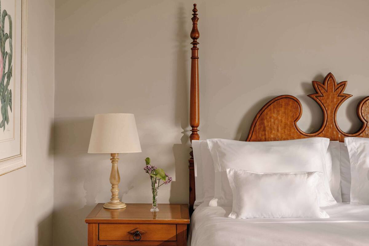A neatly made bed with white linens, a wooden nightstand with a lamp and a vase of flowers, and a framed picture on the wall.