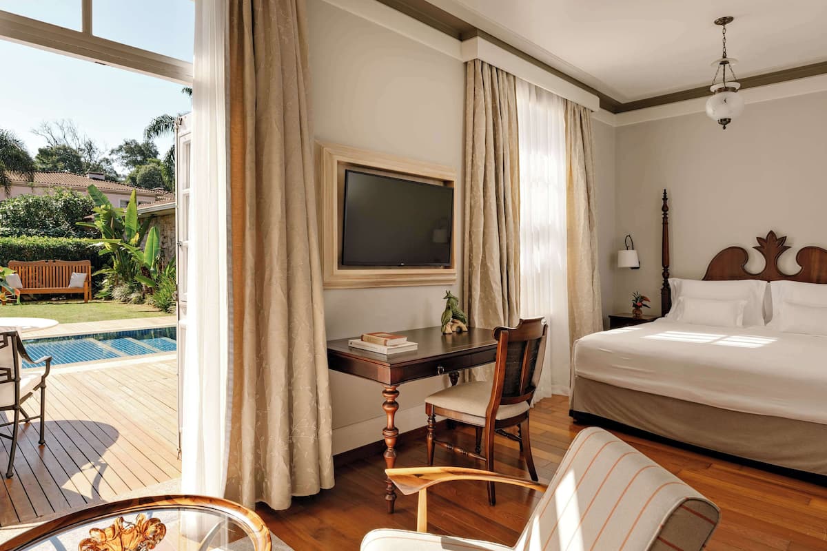 A hotel room with a bed, a desk with a chair, a TV, and a view of a pool and garden.