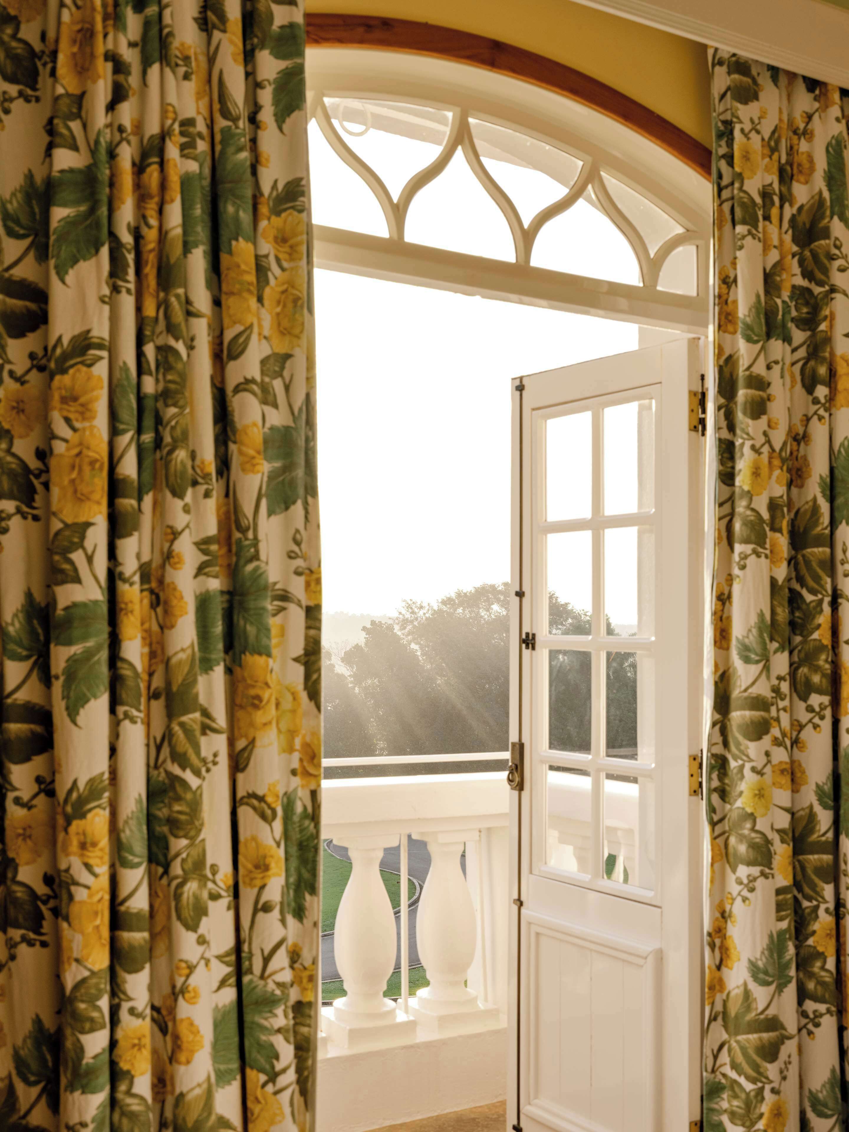 Una habitación con una puerta blanca, cortinas florales y vistas al exterior con vegetación.