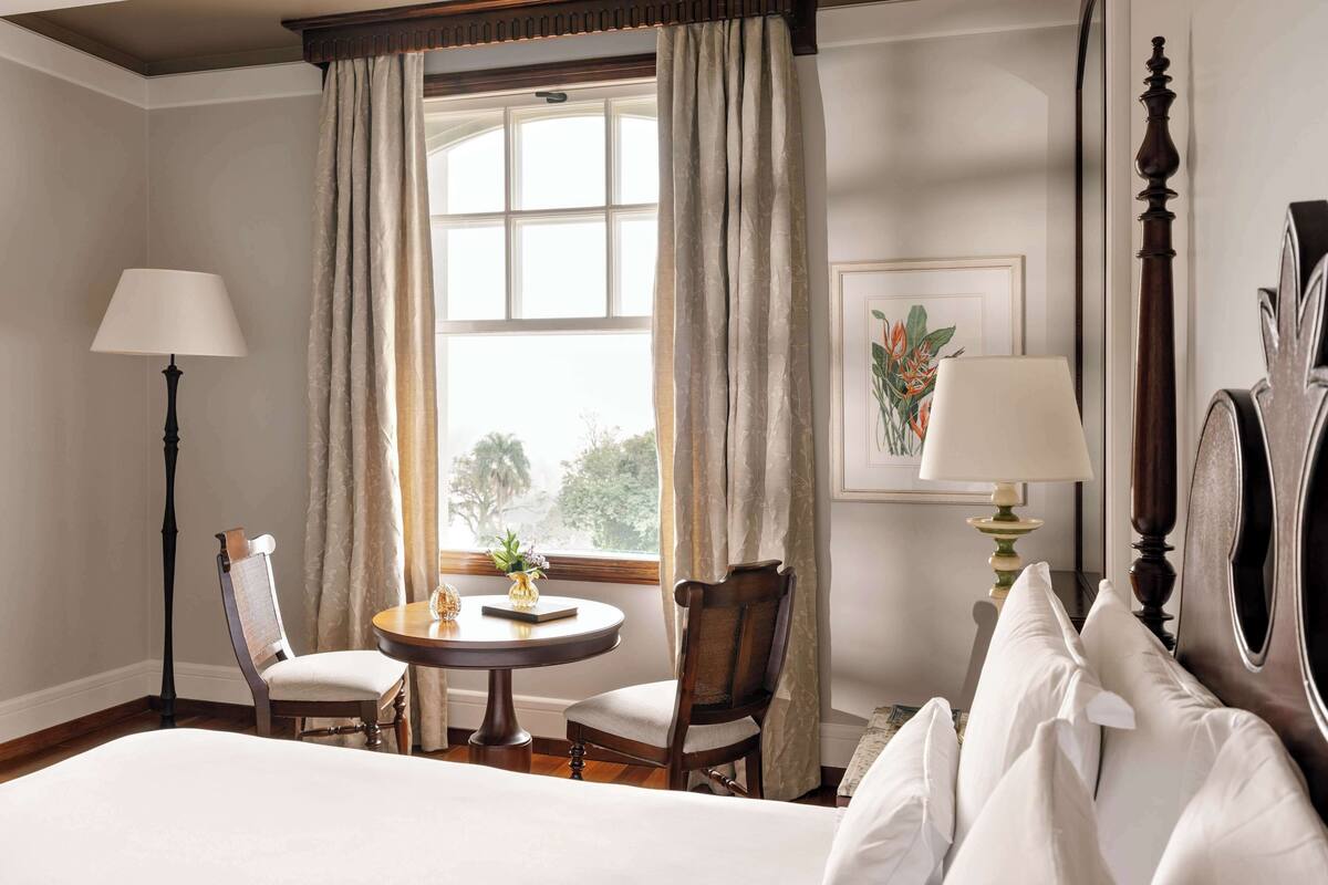 A hotel room with a bed, a round table with chairs, a desk, a lamp, and a window with curtains.