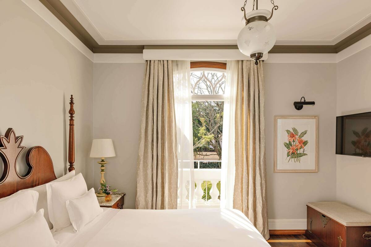 A bedroom with a large bed, a wooden headboard, a bedside table with a lamp, a window with curtains, a framed picture on the wall, and a wooden chest of drawers.