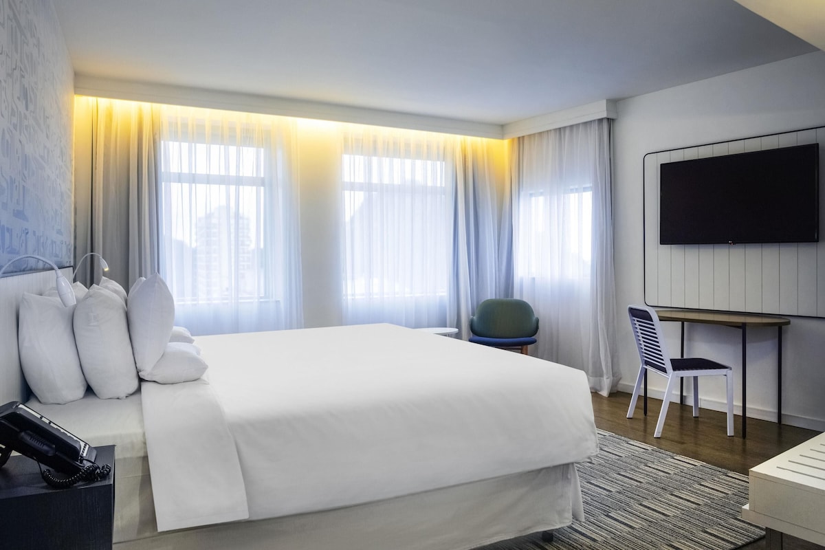A modern hotel room with a large bed, a desk, a chair, and a TV on the wall.