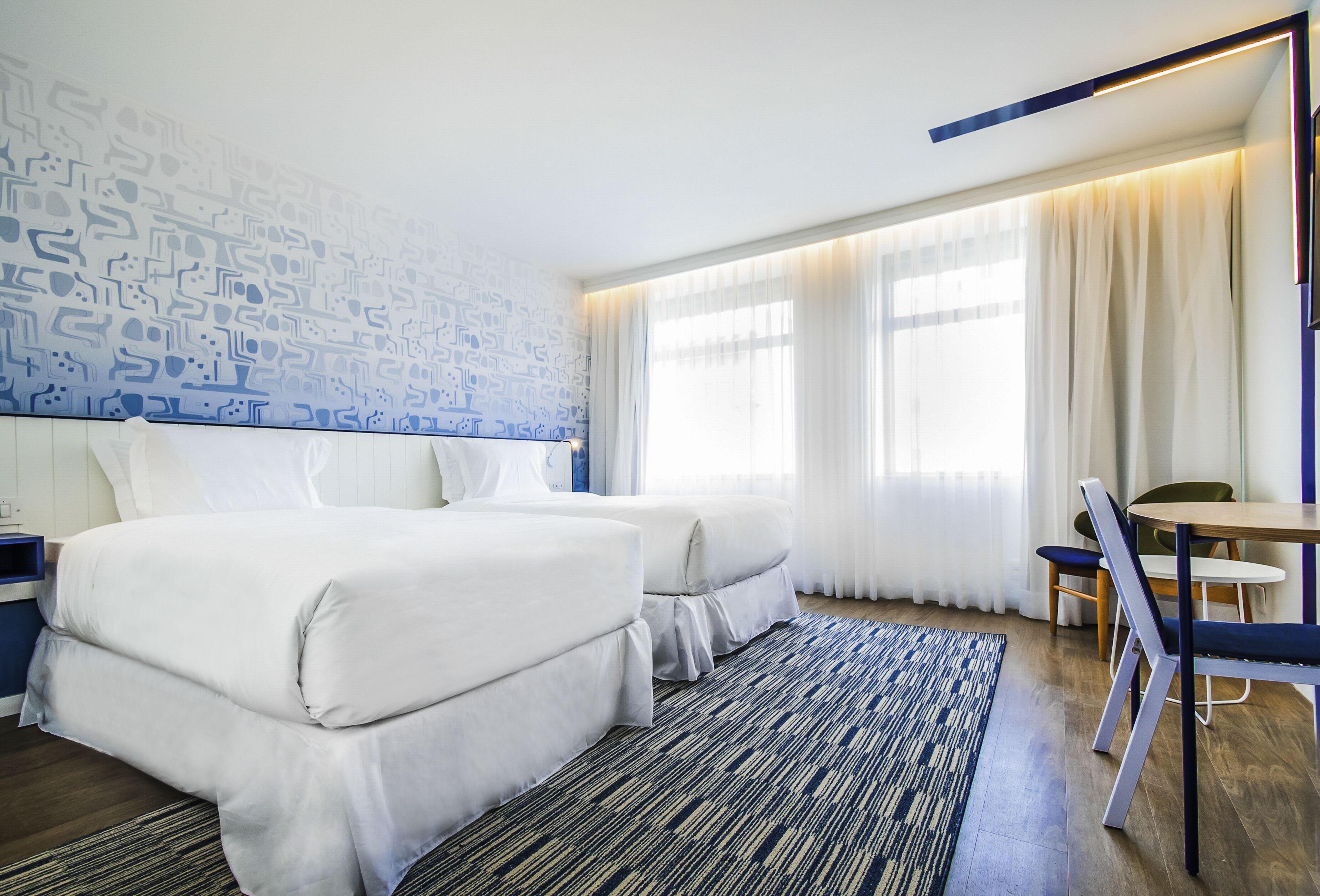 Una habitación de hotel con dos camas, un escritorio, una silla y un ventanal con cortinas.