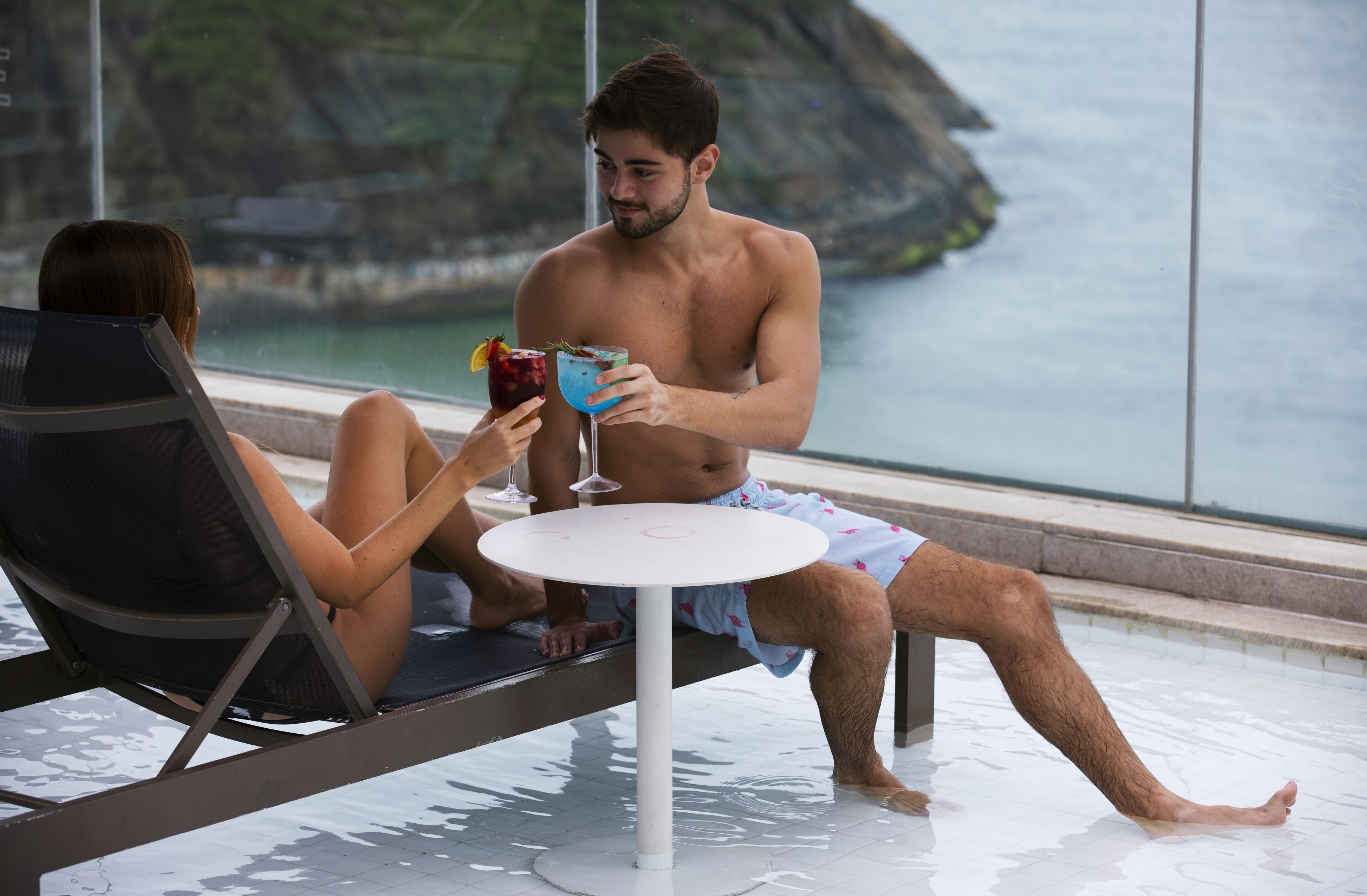 Una pareja descansando junto a una piscina con bebidas, con vistas a un cuerpo de agua y un paisaje rocoso.