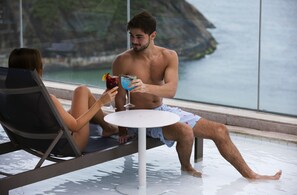 A couple relaxing by a pool with drinks, overlooking a body of water and a rocky landscape. - Novotel Rio de Janeiro Leme (Rio de Janeiro)