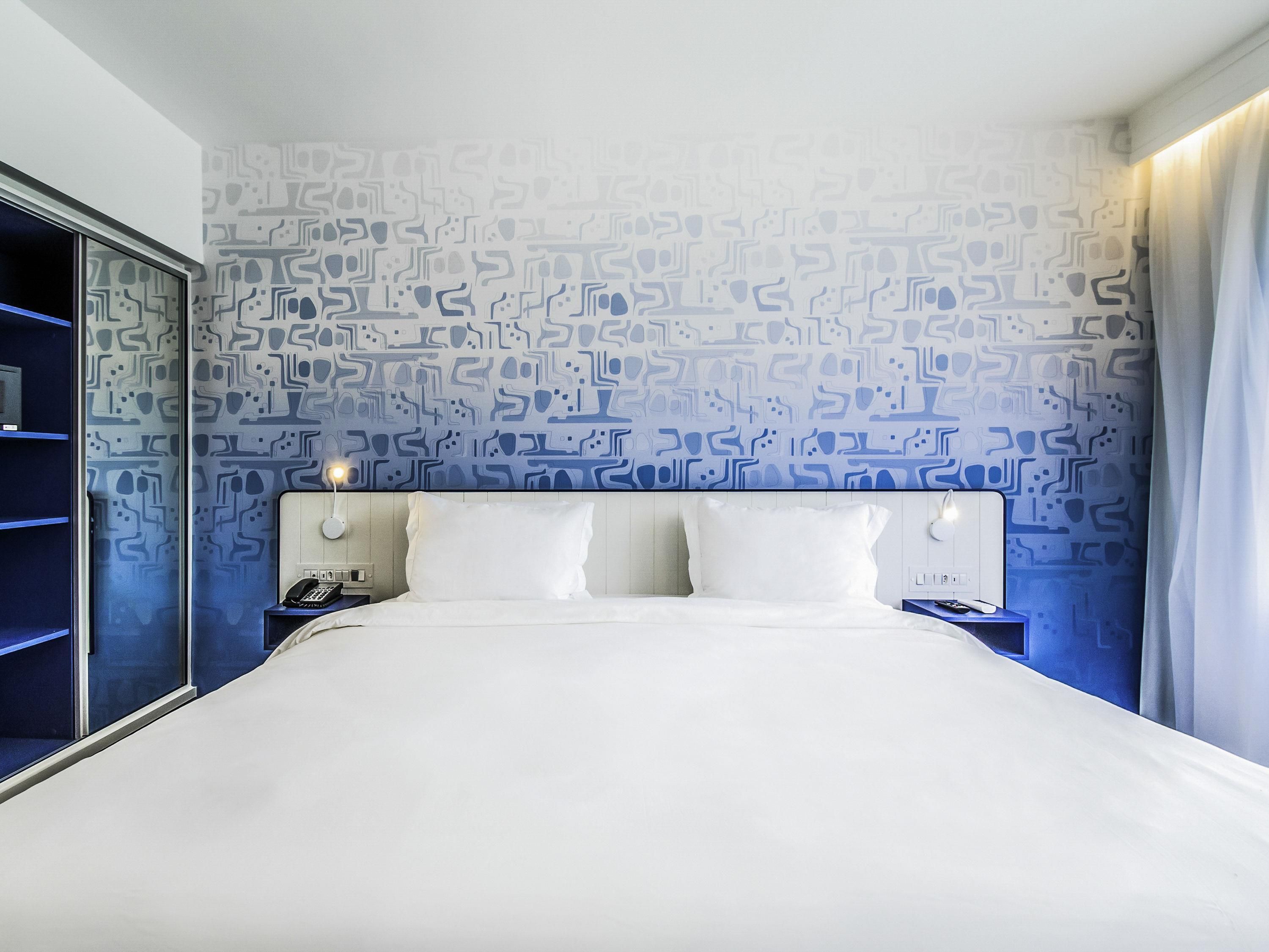 Habitación de hotel con pared estampada, cama con sábanas blancas, dos mesitas de noche con lámparas y un tocador con teléfono.
