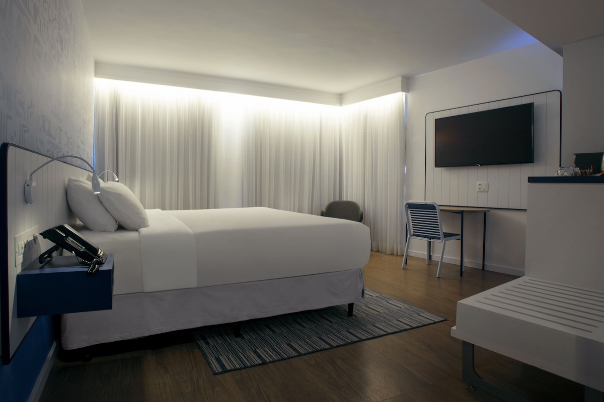 A modern hotel room with a large bed, a desk, a chair, and a flat-screen TV mounted on the wall.