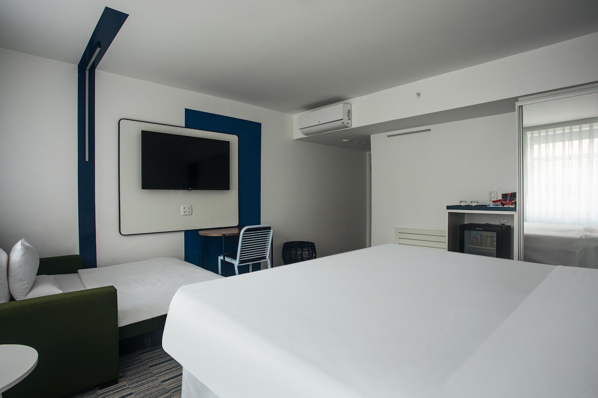 A hotel room with a large bed, a TV mounted on the wall, a desk with a chair, and a small kitchenette.