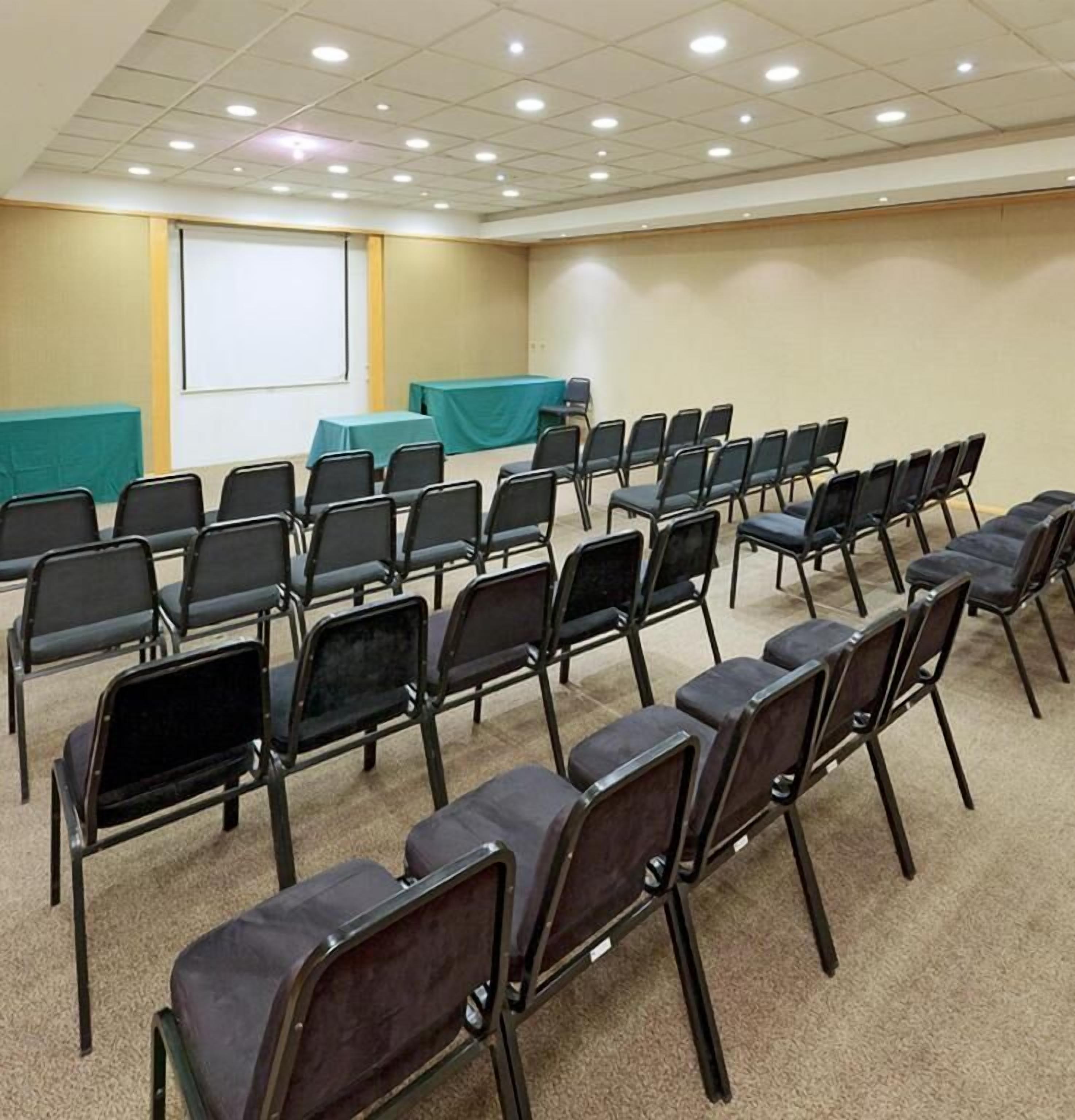 Une salle de conférence avec des rangées de chaises, un écran de projection et un sol recouvert de moquette.