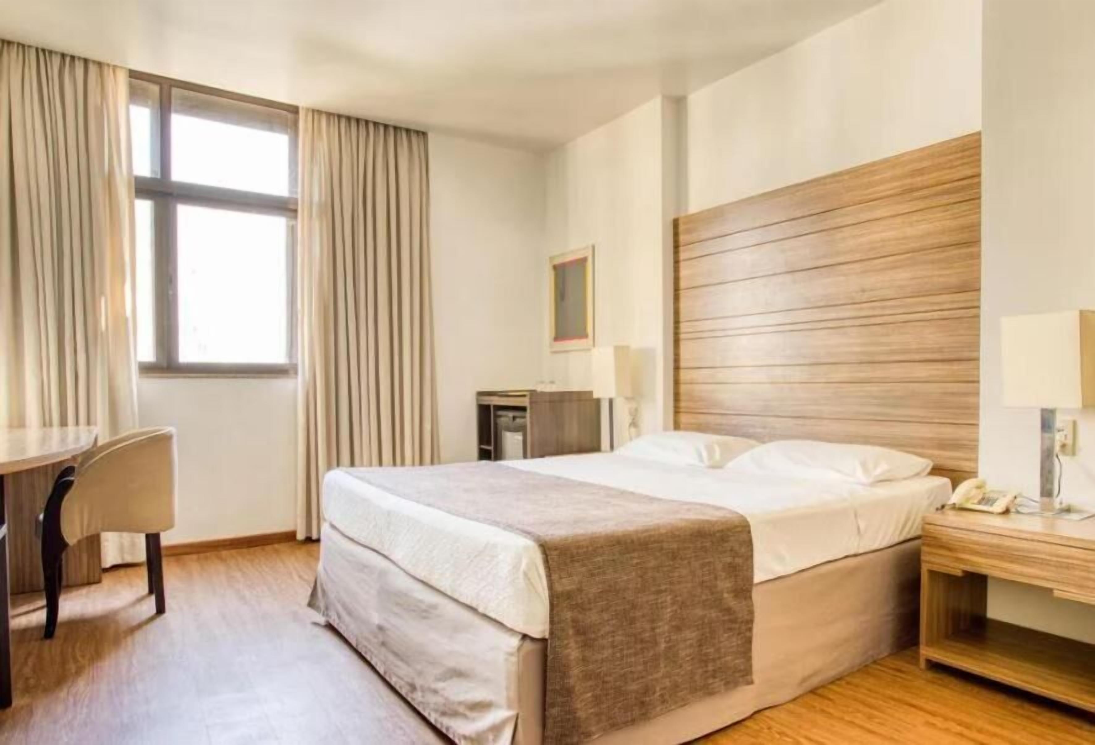 Une chambre d’hôtel comprenant un lit, une table de chevet, une lampe, un bureau, une chaise et une fenêtre avec des rideaux.