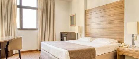 Une chambre d’hôtel comprenant un lit, une table de chevet, une lampe, un bureau, une chaise et une fenêtre avec des rideaux.