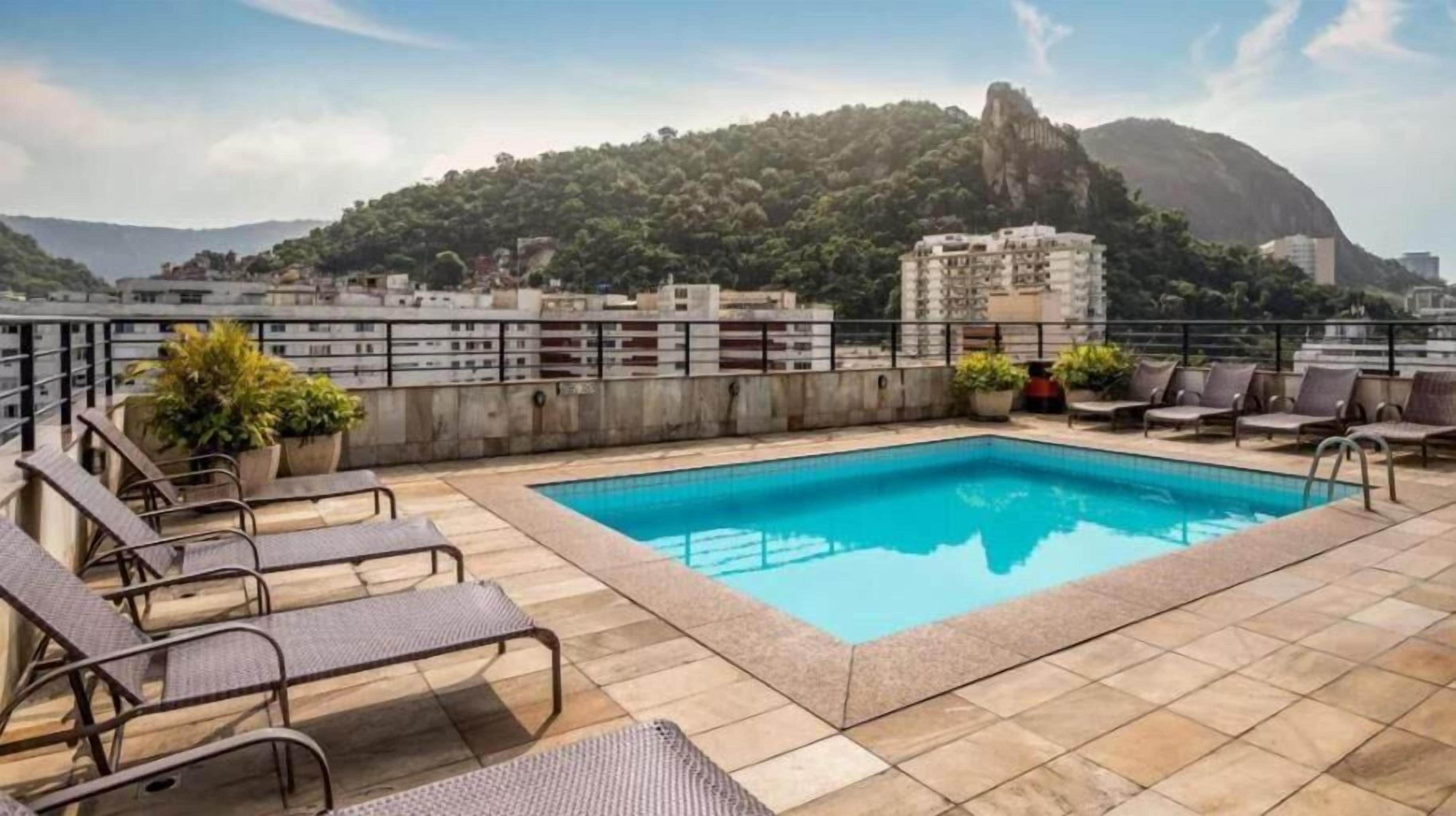 Una piscina al terrat amb gandules i plantes en testos, envoltada d'un paisatge urbà i una muntanya.