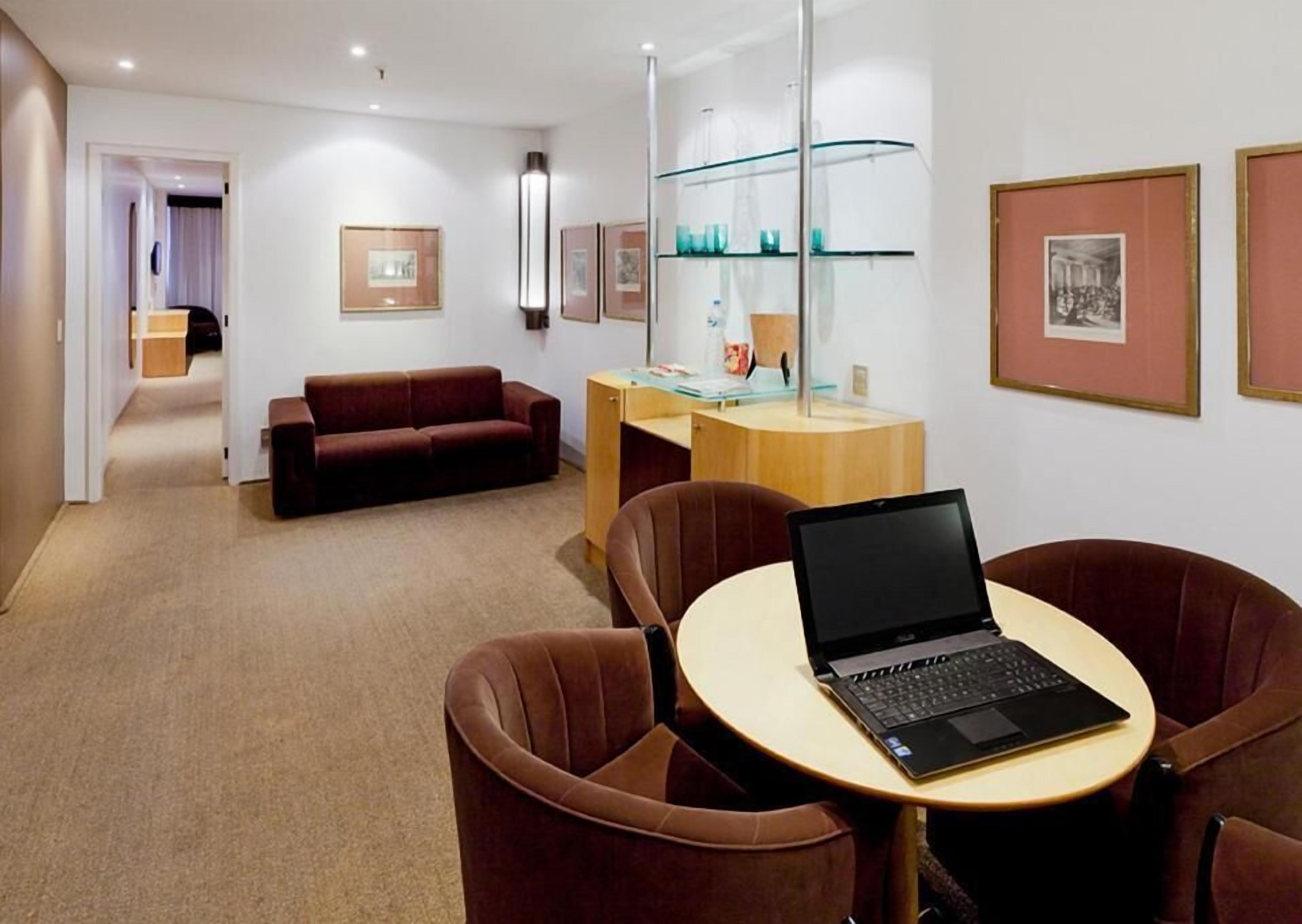A modern hotel room with a brown leather sofa, a wooden desk with a laptop, and framed pictures on the walls.