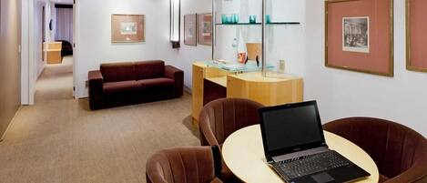 A modern hotel room with a brown leather sofa, a wooden desk with a laptop, and framed pictures on the walls.
