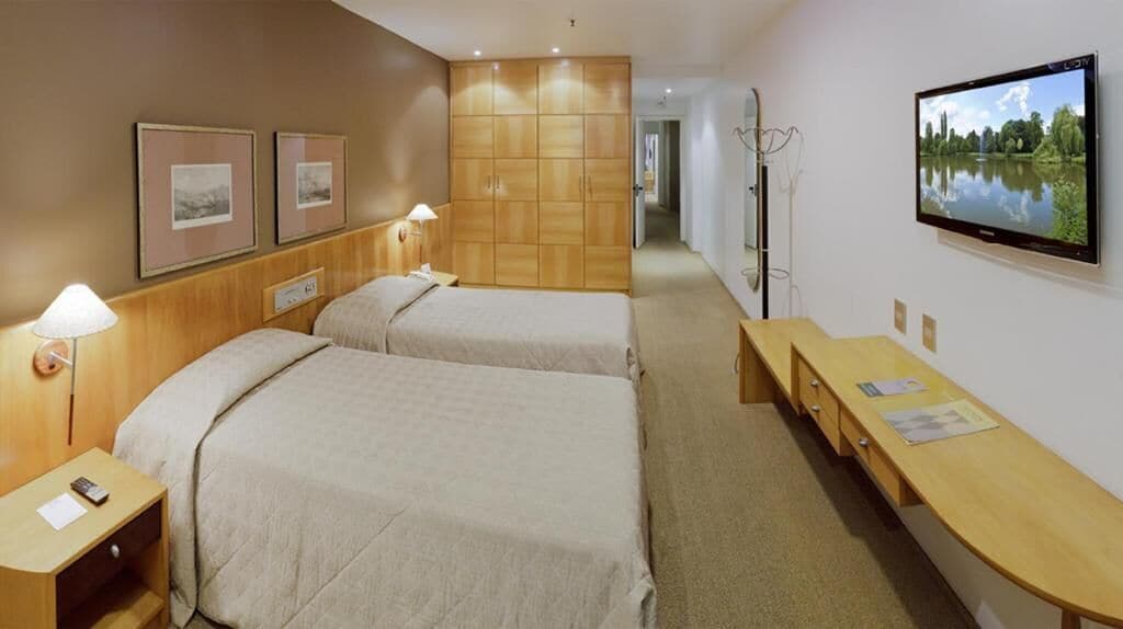 Habitación de hotel con dos camas, un armario de madera, un escritorio con papeles y un televisor mostrando un paisaje.