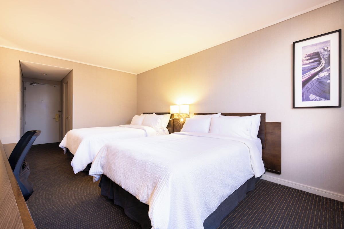 A hotel room with two beds, a desk, a chair, a lamp, and a framed picture on the wall.